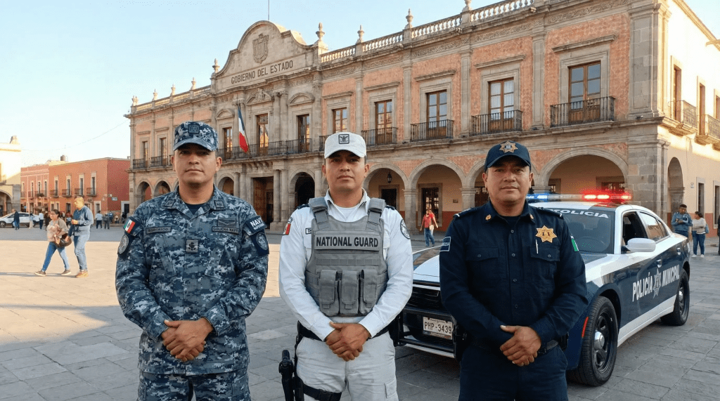 Imagen profesional y cinematográfica que representa la unificación de las fuerzas de seguridad en Tlaxcala. Tres oficiales (Marina, Guardia Nacional y Policía Municipal) posan juntos con profesionalismo frente a una patrulla moderna en la Plaza de la Constitución. La escena simboliza la confianza, la coordinación interinstitucional y el fortalecimiento de la seguridad pública bajo un enfoque de dignidad y servicio.