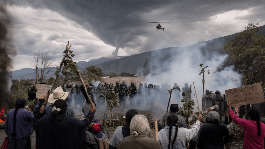 Imagen cinematográfica e hiperrealista de un enfrentamiento social en Atlangatepec, Tlaxcala. En primer plano, un grupo de comuneros, incluyendo ancianos y mujeres, se defienden con ramas y pancartas frente a una línea de policías antimotines envueltos en humo de gases lacrimógenos. Al fondo, un helicóptero sobrevuela ominosamente y nubes de tormenta con forma de tornado se ciernen sobre las montañas. La escena, con el humo de llantas quemadas, captura la tensión, la resistencia y la inminente tormenta de un conflicto social.
